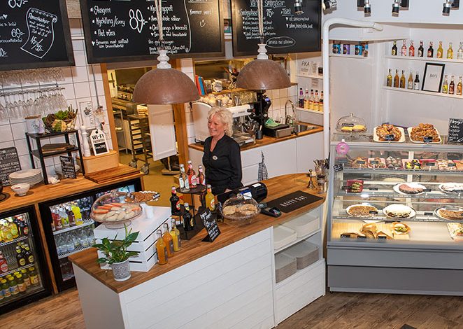 Foto i fågelperspektiv inifrån Tinas café å sånt, i kyldisken finns kakor och bakom disken står en leende kvinna
